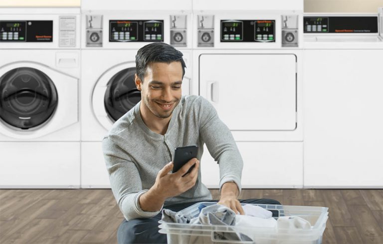 Man with laundry basket in a common area laundry room uses phone for mobile pay