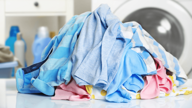 Pile of colorful clothes on a surface with a washing machine and detergent bottles in the background.