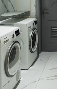 Two white front-loading laundry machines on a tiled floor near a gray door.