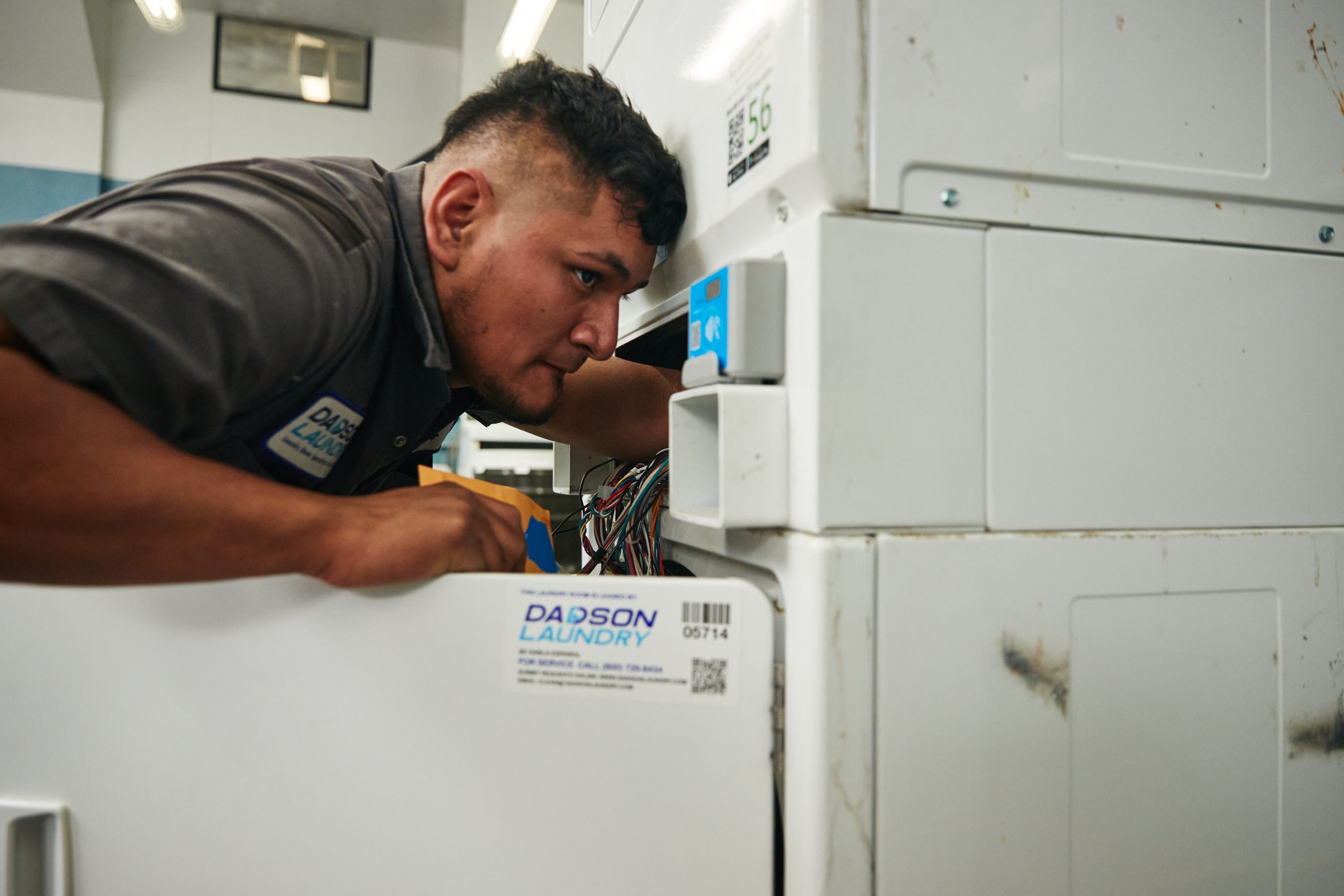 Man inspecting wiring inside an open laundry machine in a service room.