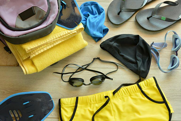 Yellow swim trunks, black swim cap, goggles, flippers, towel, blue swimwear, and sandals on wood floor.
