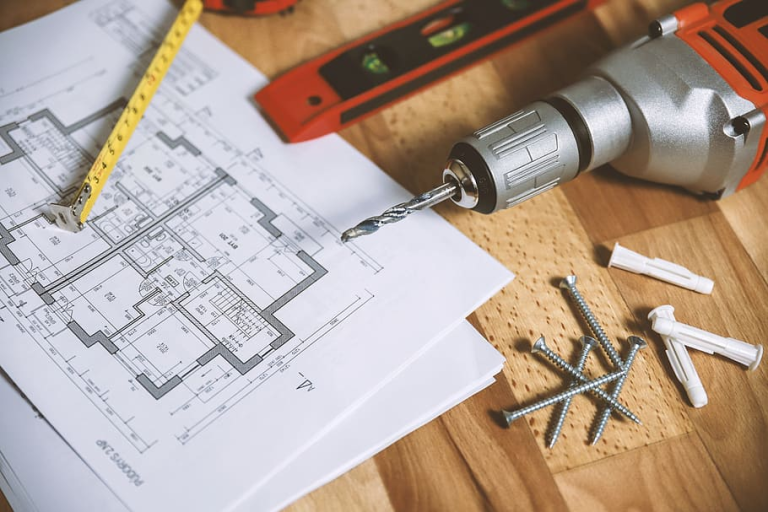 Architectural blueprints, tape measure, spirit level, power drill, screws, and wall plugs on wood surface.